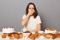 Indoor shot of overeating young adult woman with brown hair sitting at table isolated over gray background, eats lot of sweet