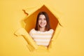 Indoor shot of playful young adult woman stands in torn paper hole, looking through breakthrough of yellow background and winking Royalty Free Stock Photo