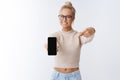Indoor shot of pleasant attractive blond woman with glasses extend smartphone to camera pointing at mobile phone screen