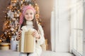 Indoor shot of pleasant looking small kid with blue charming eyes, wears santa hat, holds present in wrapped box, sits over decora