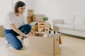 Indoor shot of pleased brunette woman dressed in white t shirt and jeans, plays with pedigree dog, moves carton box with animal,