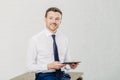 Indoor shot of pleased male owner of prosperous company dressed in formal clothes, holds modern tablet computer, enjoys online com