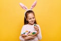 Indoor shot of positive cute little girl with rabbit ears with basket of colored eggs, looking at patters on spring symbols,