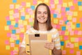 Indoor shot of positive optimistic woman student with brown hair in striped t shirt standing against yellow wall with memo cards, Royalty Free Stock Photo