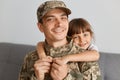 Indoor shot of positive young adult man soldier wearing camouflage uniform and cap, posing with his cute daughter, hugging each Royalty Free Stock Photo