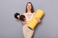 Indoor shot of pretty charming young woman wearing sportswear holding dumbbell and yoga mat, being ready to do fitness training, Royalty Free Stock Photo