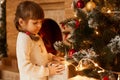 Indoor shot of pretty little girl standing near Christmas tree, keeping hands on present box, finishing to decorate Xmas tree, Royalty Free Stock Photo