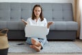 Indoor shot of puzzled helpless young adult woman wearing white shirt and jeans sitting on floor near cough and working on laptop