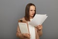 Indoor shot of sad angry aggressive brown-haired woman wearing jacket holding clipboard with documents, reading papers with