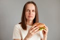 Indoor shot of sad attractive woman keeps diet, feels hungry, holding american hamburger in hands, looking at camera with sorrow,