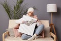 Indoor shot of sad young adult woman in coat and hat sit in cold living room holding electric heater, struggling from chill freeze Royalty Free Stock Photo