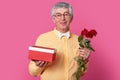 Indoor shot of satisfied elderly gentleman with satisfied expression holds gift box and red roses, dressed in yellow shirt with