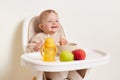 Indoor shot of satisfied little female girl wearing beige sweater sitting in high chair and eating, posing isolated over white