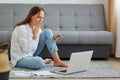 Indoor shot of scared woman wearing white shirt and jeans, sitting on the floor with laptop computer, looking at screen of Royalty Free Stock Photo