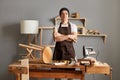 Indoor shot of serious concentrated craftsman standing in workshop and looking at camera, wearing brown apron and black cap, keeps