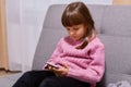 Indoor shot of serious concentrated dark haired little girl wearing warm pink sweater, holding mobile phone in hands and playing