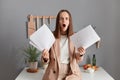 Indoor shot of shocked amazed surprised young adult woman wearing beige suit, holding paper documents, having troubles with