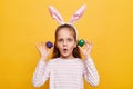 Indoor shot of shocked surprised girl with rabbit ears on her head with eggs isolated on yellow background, looking at camera with Royalty Free Stock Photo