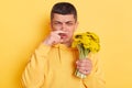 Indoor shot of sick ill man wearing casual style hoodie posing isolated over yellow background, holding bouquet of dandelions and Royalty Free Stock Photo