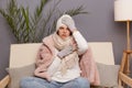 Indoor shot of sick unhealthy frozen woman wearing cap, gloves, scarf and coat sitting in living room, fells terrible headache and Royalty Free Stock Photo