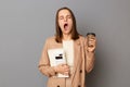 Indoor shot of sleepy tired woman wearing beige jacket standing with organizer and calculator in hands isolated over gray Royalty Free Stock Photo