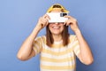 Indoor shot of smiling delighted teenager girl wearing baseball cap and stripe T-shirt, covering her eyes with mobile phone,