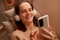 Indoor shot of smiling girl relaxing at home, listening to music using smartphone and wearing white headphones, looking at cell Royalty Free Stock Photo