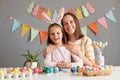 Indoor shot of smiling happy mother and her charming dark haired daughter posing among painted Easter eggs, child wearing bunny