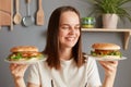 Indoor shot of smiling happy satisfied woman with dark hair dressed in white T-shirt holding two big burger sandwiches, being Royalty Free Stock Photo