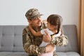 Indoor shot of smiling man soldier wearing camouflage uniform and cap, posing with his daughter, hugging each other and being Royalty Free Stock Photo