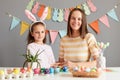 Indoor shot of smiling mother and daughter wearing bunny ears painting eggs preparing for Easter against gray decorated wall, Royalty Free Stock Photo