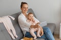 Indoor shot of smiling positive dark haired woman wearing white shirt and jeans sitting on sofa with her baby daughter, having Royalty Free Stock Photo