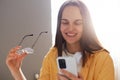 Indoor shot of smiling woman with brown hair wearing yellow shirt posing in office, holding her glasses and using cell phone, Royalty Free Stock Photo