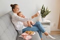 Indoor shot of smiling woman wearing white shirt and jeans sitting on sofa with baby daughter, female holding smart phone and Royalty Free Stock Photo