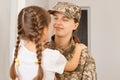 Indoor shot of smiling young adult woman wearing camouflage uniform and cap hugging and looking in eyes to her little daughter Royalty Free Stock Photo
