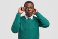 Indoor shot of stupefied black man with amazed facial expression, keeps hand on rim of spectacles, dressed in casual