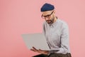 Indoor shot of successful copywriter types publication, reads information, holds laptop computer, wears glasses, shirt, headgear,