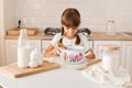 Indoor shot of surprised charming little girl baker on kitchen with baking ingredients mixing dough in pot, bakes pie or cake,
