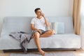 Indoor shot of thoughtful Caucasian brunette man wearing white shirt and jeans, sitting on gray couch in light living room, Royalty Free Stock Photo