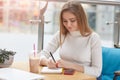 Indoor shot of thoughtful female student prepares for sesssion in cafeteria, writes down some inforamtion, drinks fresh cold