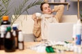 Indoor shot of tired exhausted bored woman sitting on sofa wrapped in scarf surrounded with medicines, drinking tea, holding cup, Royalty Free Stock Photo