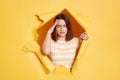 Indoor shot of tired exhausted young adult brunette woman stands in torn paper hole, suffering headache, touching head, looking Royalty Free Stock Photo