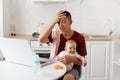 Indoor shot of tired handsome freelancer male wearing burgundy t shirt, posing in white kitchen, keeping hand on forehead, feels