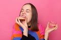 Indoor shot of very hungry woman with brown hair wearing sweater biting sugary tasty donut, keeps eyes closed, enjoying delicious