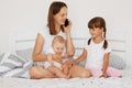 Indoor shot of winsome woman wearing white casual style t shirt sitting on bed with her children and talking via cell phone, Royalty Free Stock Photo