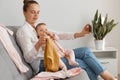 Indoor shot of woman blogger wearing white shirt and jeans sitting on sofa with baby daughter and having livestream, showing kid` Royalty Free Stock Photo