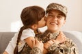Indoor shot of woman soldier returning home from army, wearing camouflage uniform and cap, charming daughter hugging and kissing Royalty Free Stock Photo