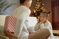 Indoor shot of woman wearing white jumper posing backwards giving present box to her daughter while sitting near Christmas tree, Royalty Free Stock Photo