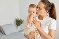Indoor shot of young adult attractive woman holding daughter wrapped in towel after bathroom, mother kissing her lovely baby girl Royalty Free Stock Photo