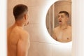 Indoor shot of young adult brunette male with naked upper body showing tongue in front of mirror in bathroom, doing morning